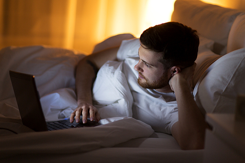 technology, internet, communication and people concept - young man with laptop computer in bed at home bedroom at night