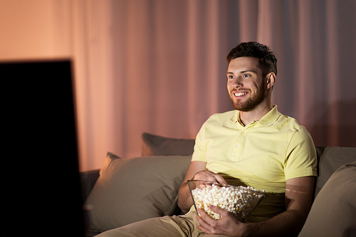 people, mass media, television and entertainment concept - happy smiling young man watching tv and eating popcorn at night at home