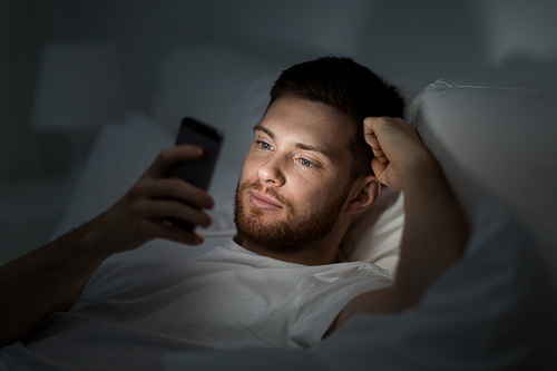 technology, internet, communication and people concept - young man texting on smartphone in bed at home at night