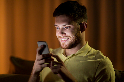 technology, internet, communication and people concept - happy smiling young man texting on smartphone at home at night