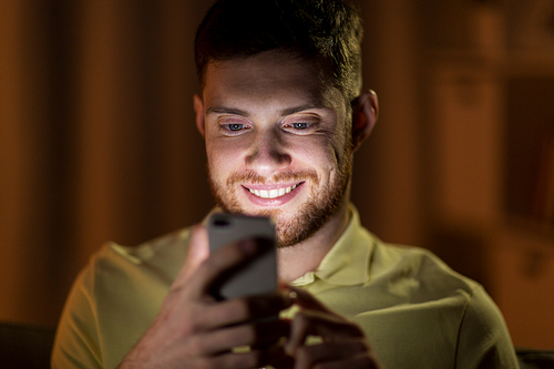 technology, internet, communication and people concept - happy smiling young man texting on smartphone in bed at home at night