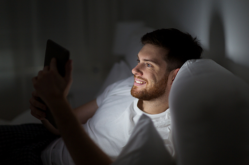 technology, internet, communication and people concept - happy young man with tablet pc computer in bed at home bedroom at night