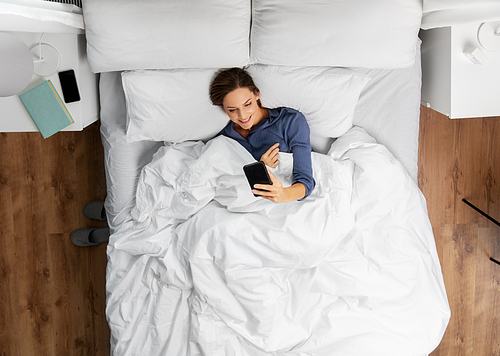 technology, internet, communication and people concept - happy young woman texting on smartphone lying in bed at home in morning