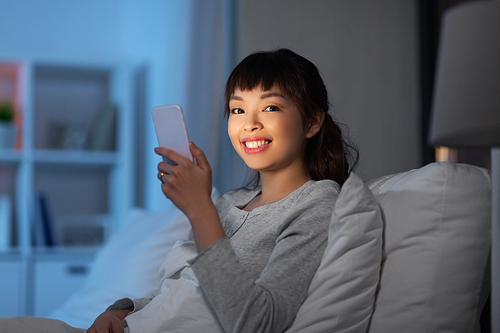 technology, internet, communication and people concept - happy smiling young asian woman with smartphone lying in bed at home at night