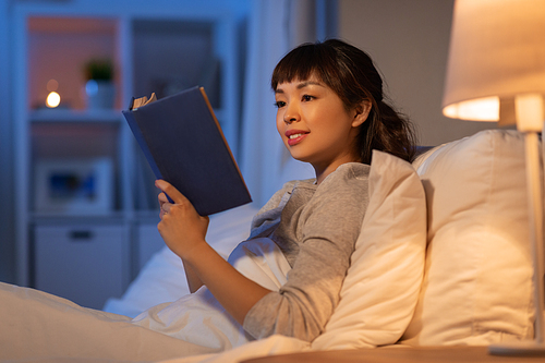 people, bedtime and rest concept - young asian woman reading book in bed at home at night
