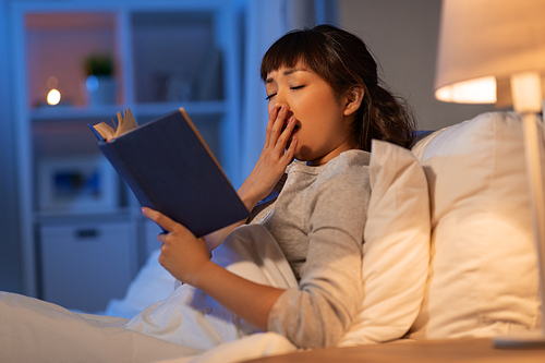 people, bedtime and rest concept - tired young asian woman reading book yawning in bed at home at night