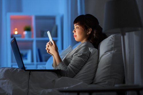 technology, internet, communication and people concept - young asian woman with smartphone and laptop lying in bed at home at night