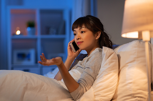 technology, communication and people concept - confused young asian woman calling on smartphone in bed at home at night