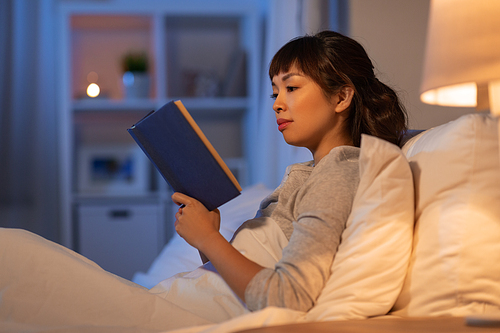 people, bedtime and rest concept - young asian woman reading book in bed at home at night