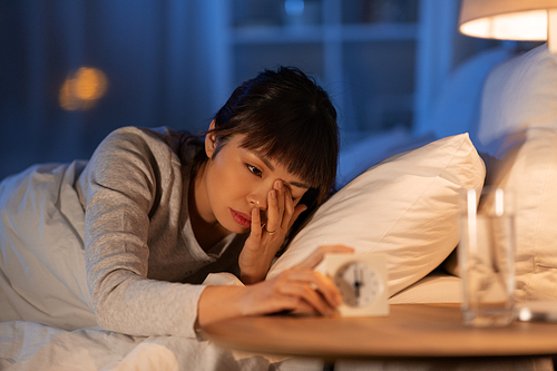 people, bedtime and rest concept - sleepy asian woman with clock lying in bed at night