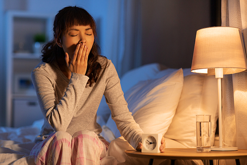 people, bedtime and rest concept - sleepy asian woman with clock yawning in bed at night