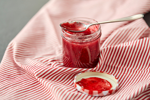 food, preserves and eating concept - mason jar with red raspberry jam and spoon on towel
