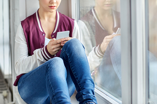 sadness and people concept - sad teenage girl with smartphone sitting on window sill