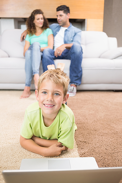 Happy little boy using laptop on the rug with parents sitting sofa