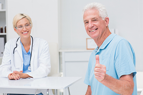 Portrait of happy senior patient gesturing thumbs up while doctor looking at him in clinic