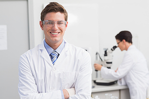 Scientist smiling at camera arms crossed and another working with microscope
