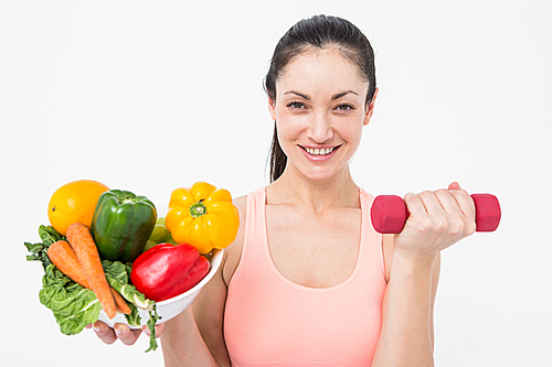 Fit brunette lifting dumbbell and holding bowl of salad on white background