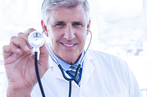 Happy doctor smiling at camera and showing his stethoscope in medical office