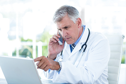 Serious doctor working on laptop and having phone call in medical office