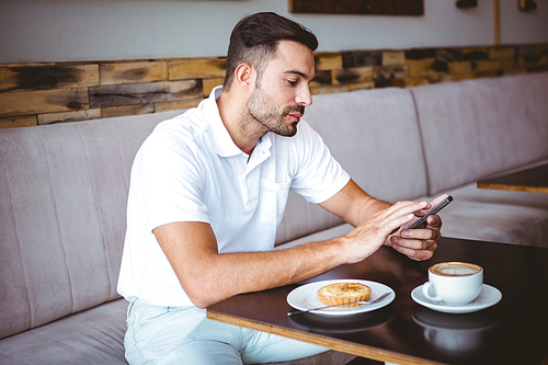 Young man having cup of coffee and pastry while texting in a cafe
