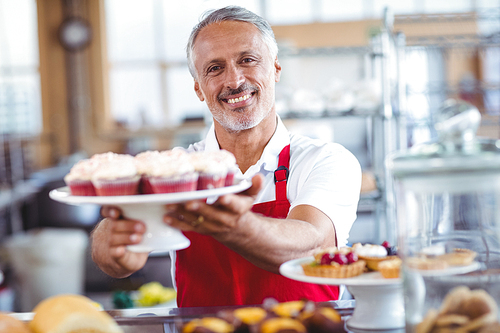 Happy barista smiling at camera and holding a plate of cupcakes in the bakery