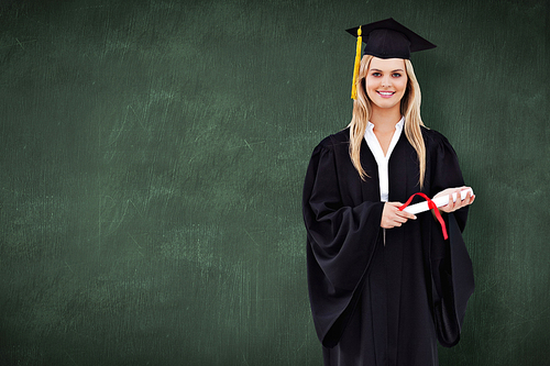Smiling blonde student in graduate robe holding her diploma against green chalkboard