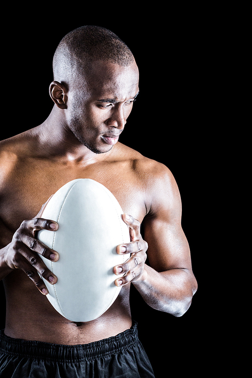 Concentrated shirtless athlete with rugby ball standing against black background