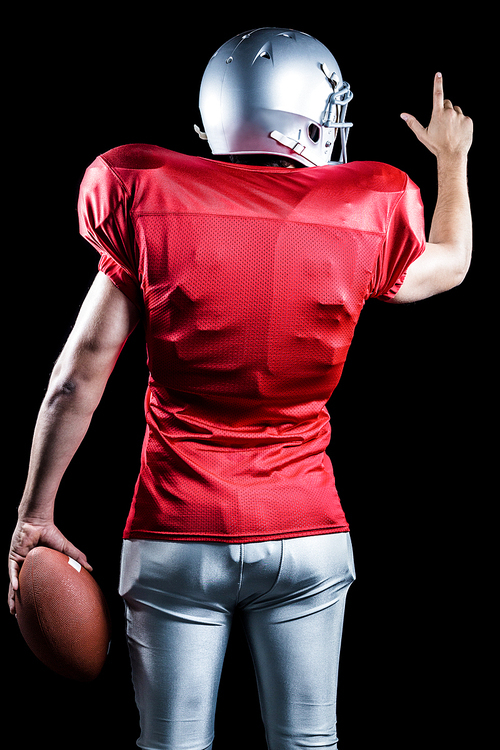 Rear view of American football player pointing while holding ball against black background