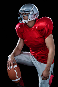 Sportsman holding American football while kneeling against black background