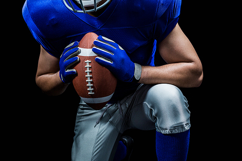 Mid section of American football player kneeling while holding ball against black background
