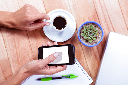 Overhead of feminine hands holding smartphone and black espresso with notebook and laptop on table