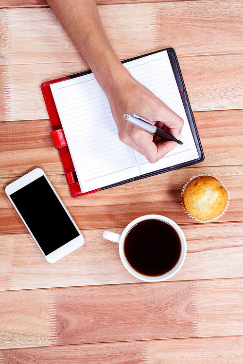 Overhead of feminine hands writing on agenda with coffee, smartphone and muffin on table