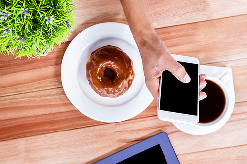 Overhead of feminine hands using smartphone with tablet, coffee and donut on table