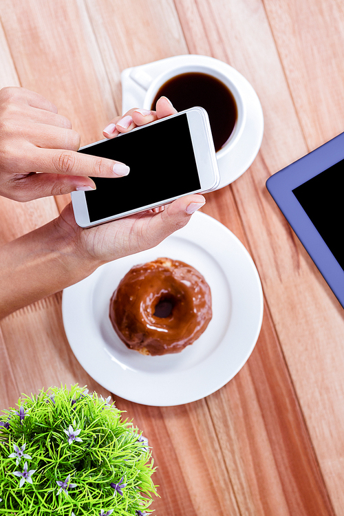 Overhead of feminine hands using smartphone with tablet, coffee and donut on table