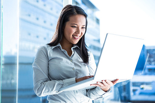 Smiling businesswoman using laptop standing near the window in office