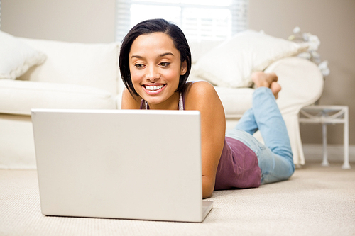 Smiling brunette using laptop lying on the carpet