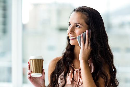 Smiling woman on phone call holding disposable cup against window