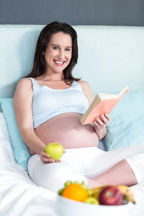 Pregnant woman eating and reading book at home