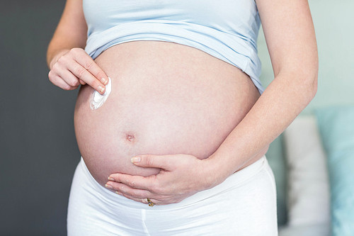 Pregnant woman applying cream on her belly in her bedroom