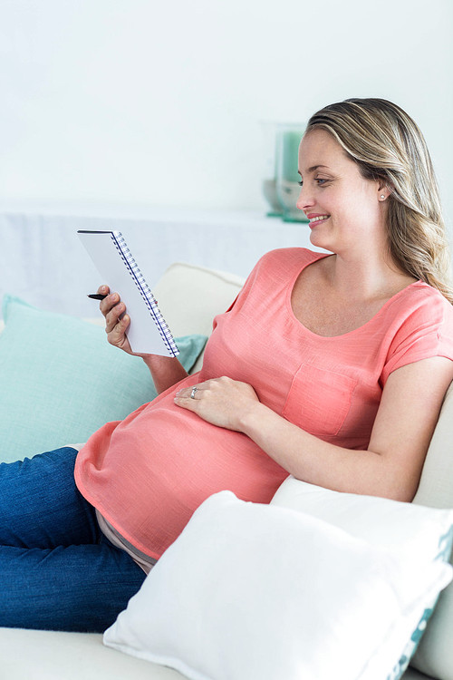 Pregnant woman writing on notepad on the couch