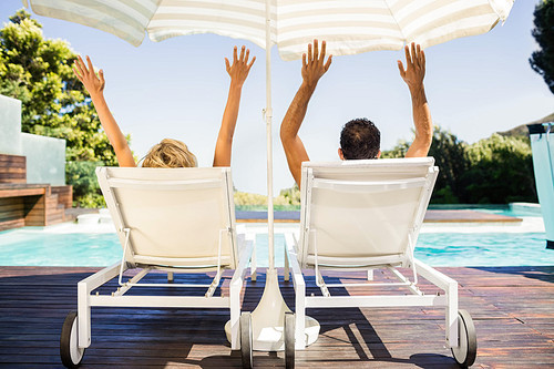 Rear view of couple raising hands and lying on deck chairs poolside