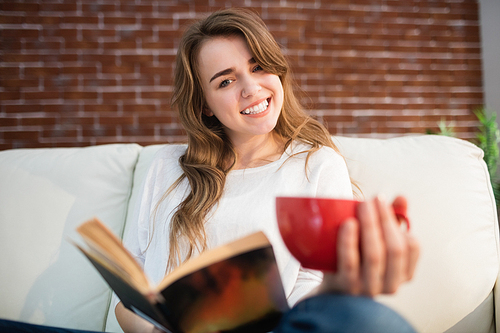 Smiling woman reading a book while drinking in the living room