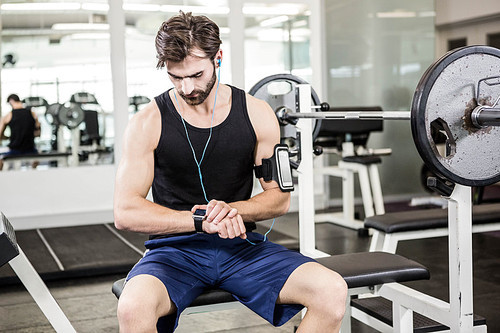Muscular man sitting on barbell bench and using smartwatch at the gym