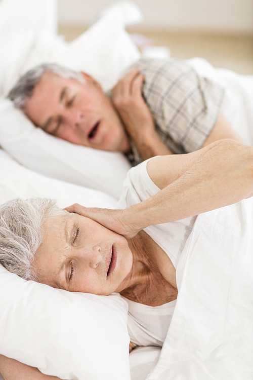 Awake senior woman in bed covering her ears while her husband is snoring