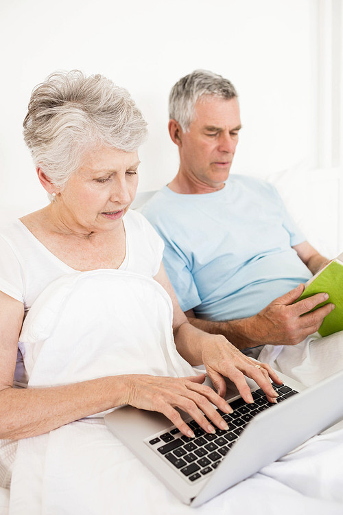 Relaxed senior couple at bed using laptop and reading boook