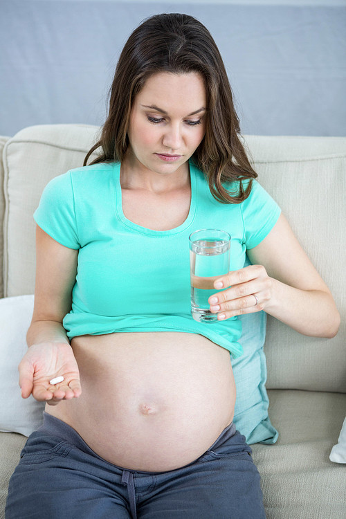 Pregnant woman holding glass of water and pills on couch