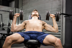Shirtless man lifting heavy dumbbells on bench at the crossfit gym