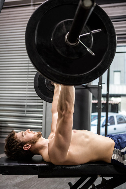Shirtless man lifting heavy barbell on bench at the crossfit gym