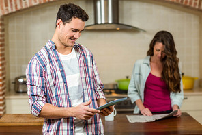 Man using tablet in kitchen while woman reading newspaper in background