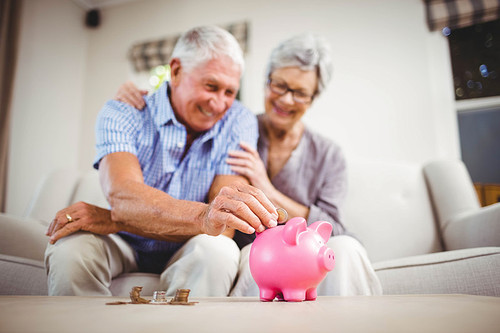 Senior man sitting with woman on sofa and putting coins in piggy bank